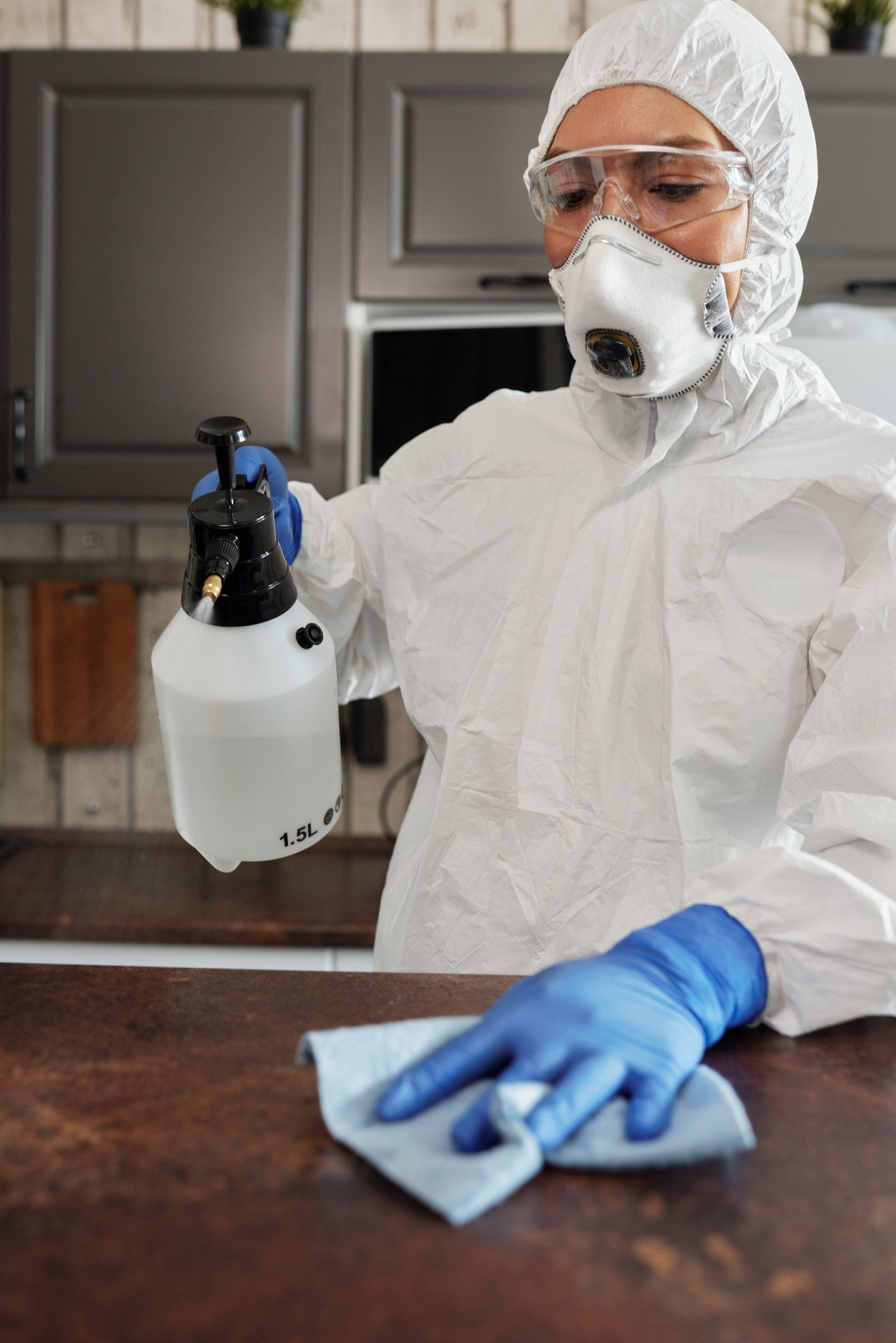 Une personne portant un équipement de protection pulvérise un produit nettoyant et essuie un comptoir de cuisine avec un chiffon bleu.