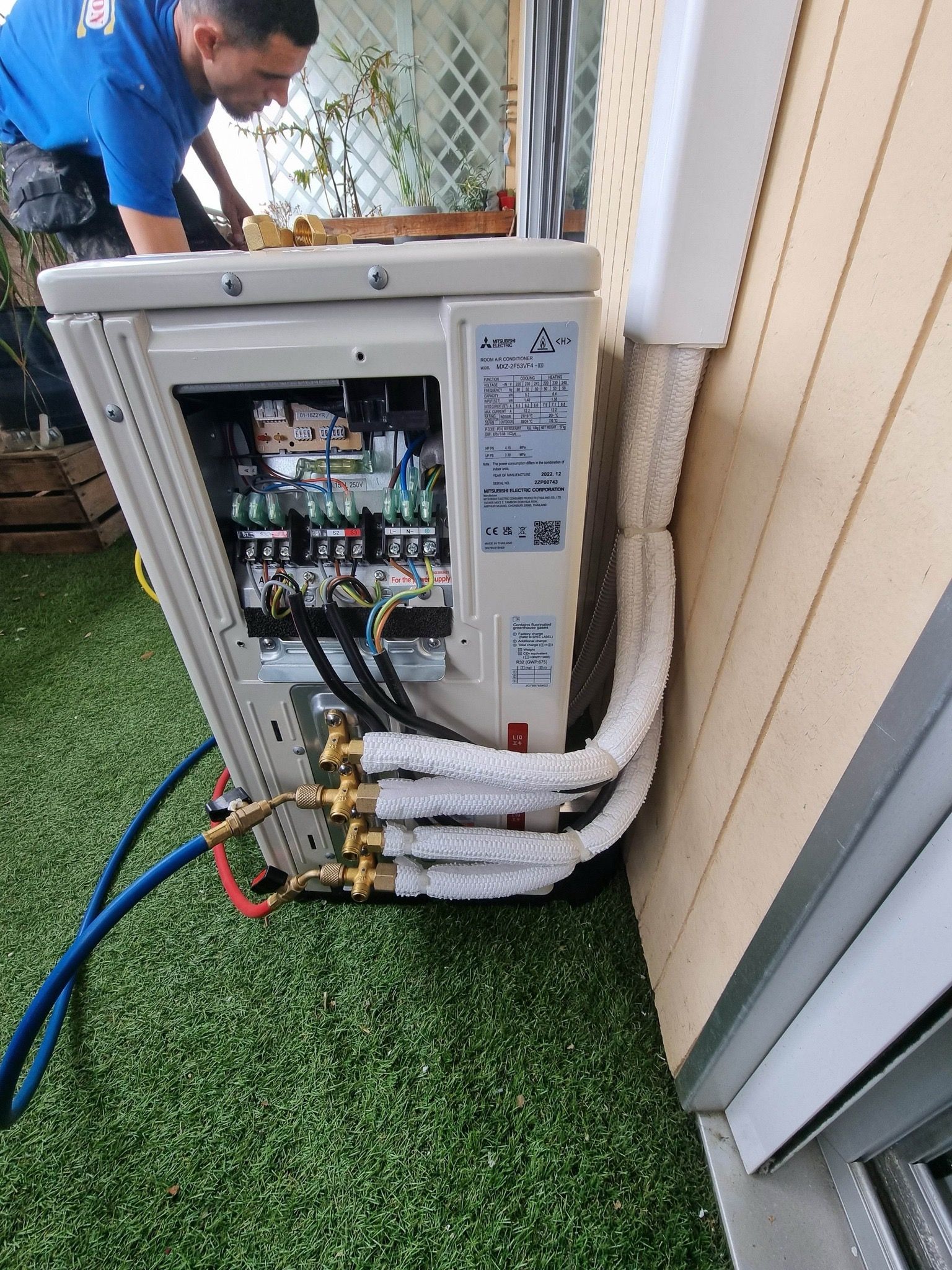 Technicien travaillant sur un tableau électrique extérieur ouvert, avec des câbles et des tuyaux apparents.