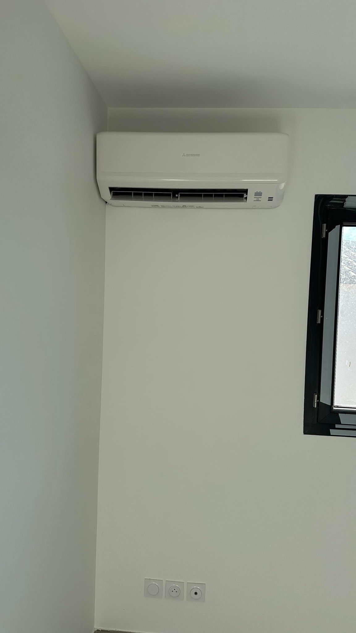 Climatiseur mural en haut d'un mur, avec des prises électriques en dessous et une fenêtre.