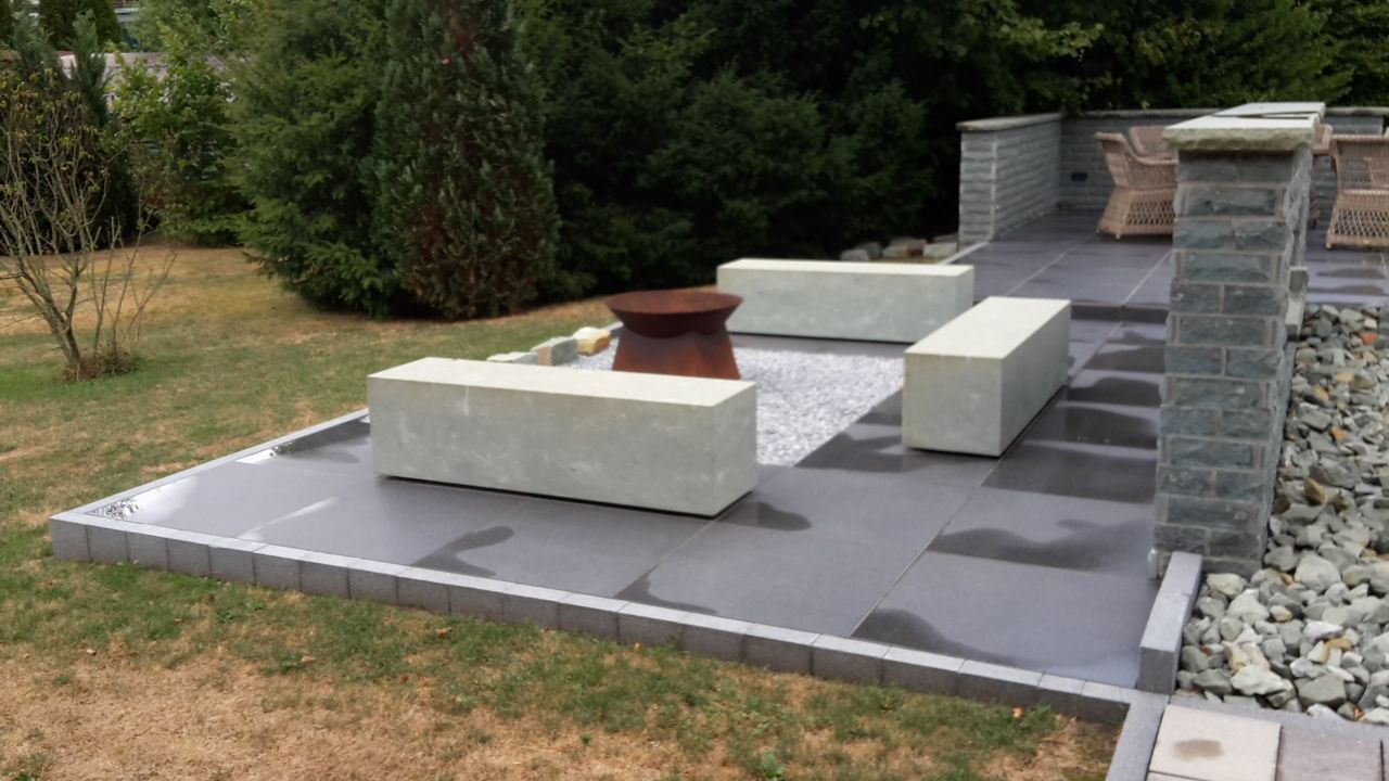 Eine mit Steinplatten besetzte Terrasse im Freien mit weißen Bänken, grauen Pflastersteinen und einem Kiesrand