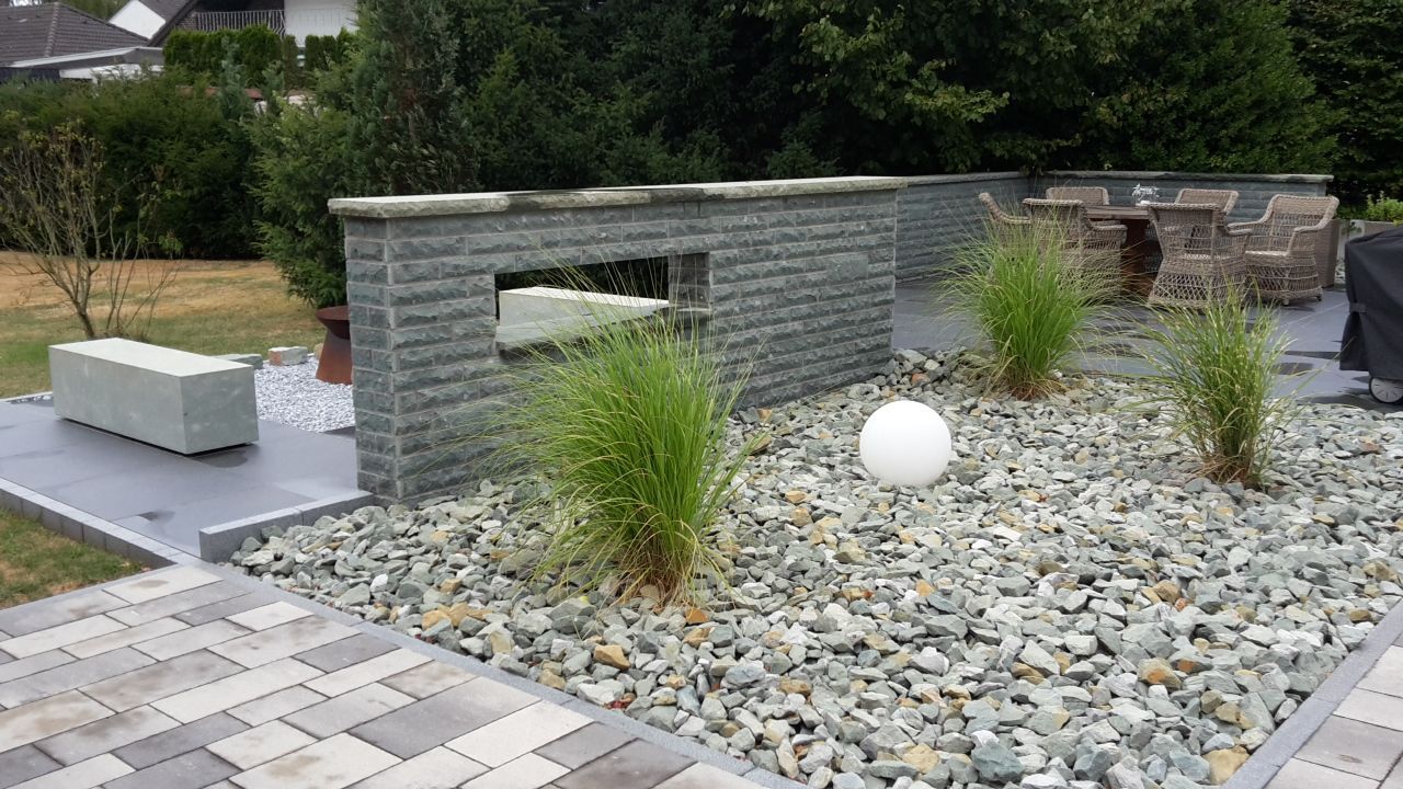Moderner Steingarten mit Steinbank, Ziergräsern und weißen Kugeln auf einer gepflasterten Terrasse