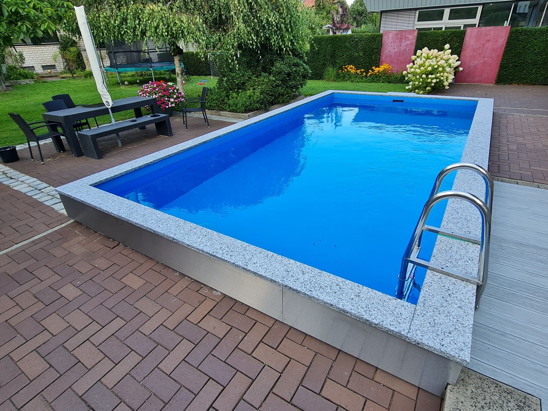 Swimmingpool im Garten mit blauem Wasser, Ziegelterrasse und Liegestühlen im Grünen
