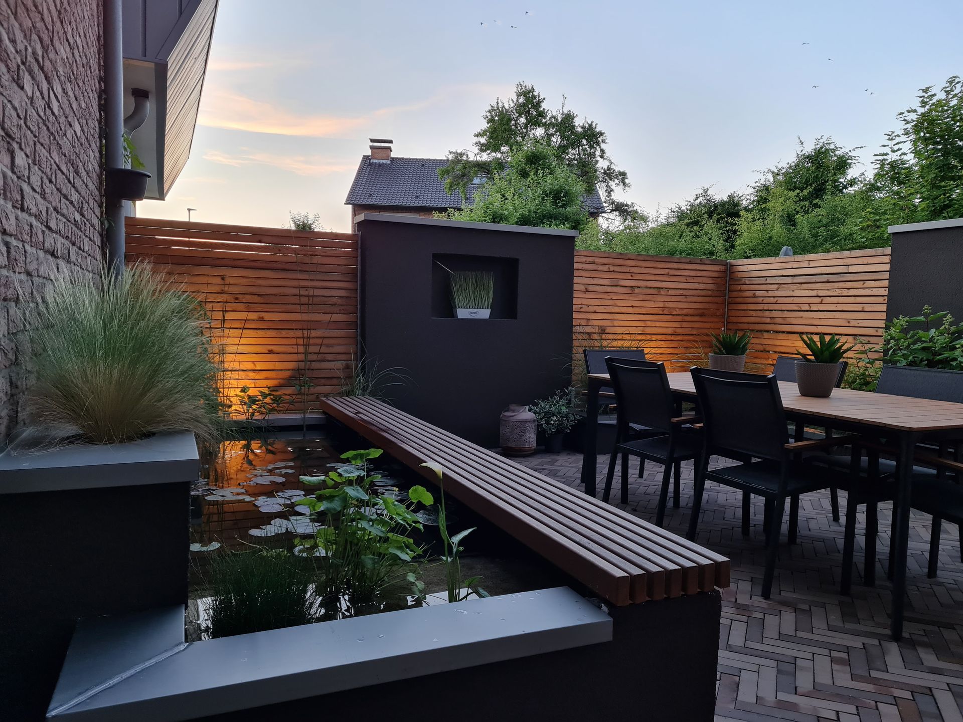 Moderne Hinterhofterrasse in der Abenddämmerung mit Holzsitzgruppen, Feuerstelle und Esstisch neben einem kleinen Teich.