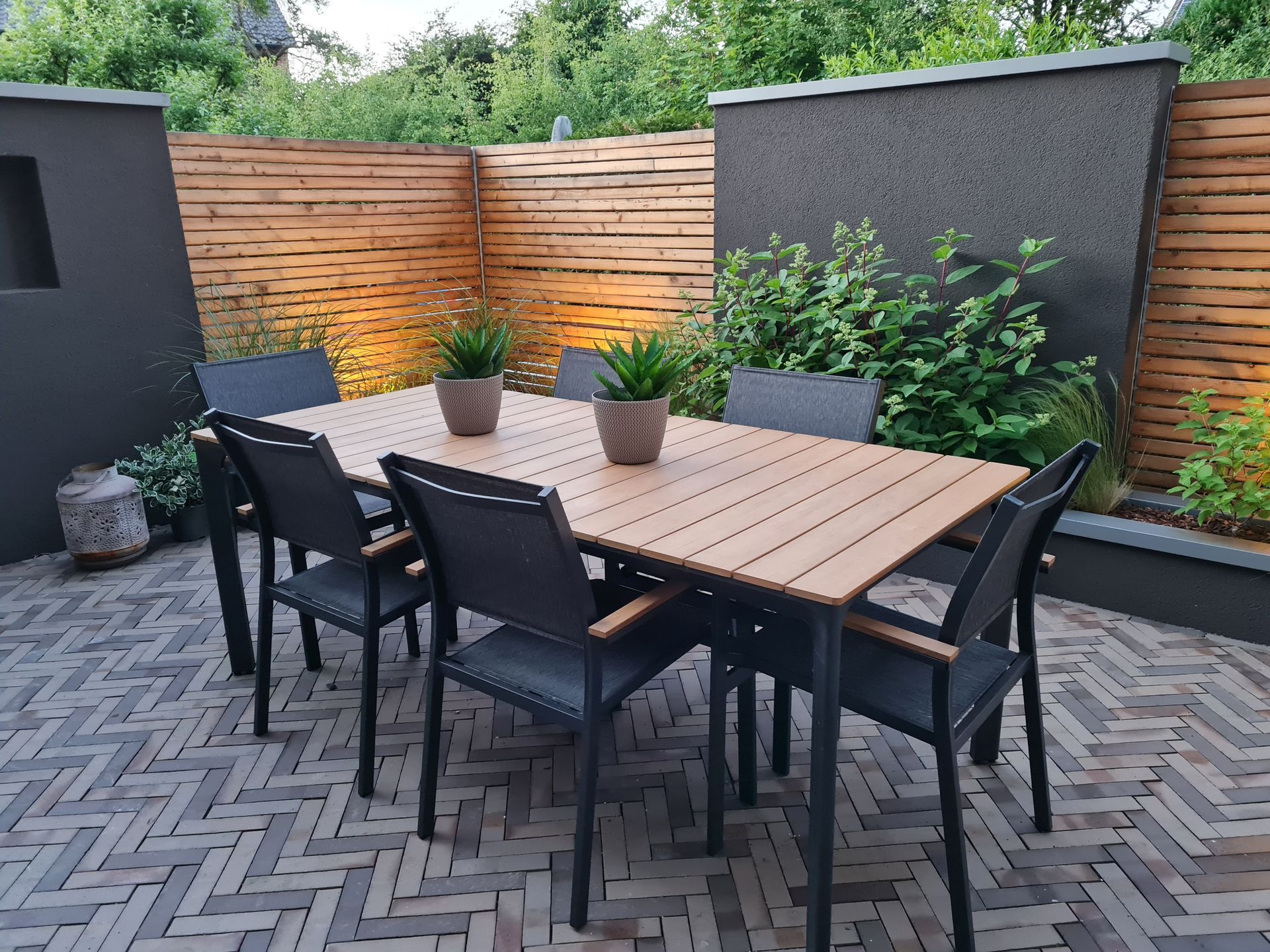 Moderne Terrasse mit schwarzen Stühlen, Holztisch, Topfpflanzen und Sichtschutzzaun in einem gepflasterten Hinterhof