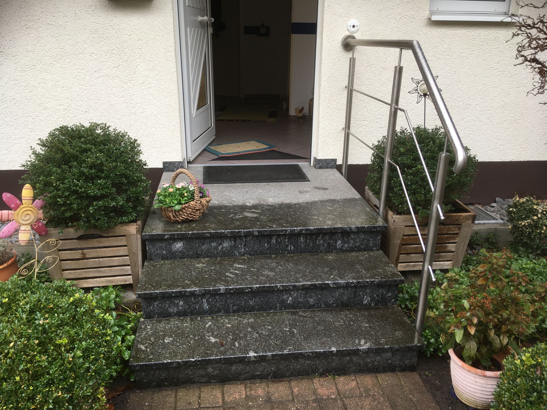 Eingangstreppe zu einem Haus mit offener Tür, Steintreppe, Metallgeländer und Topfpflanzen auf beiden Seiten.