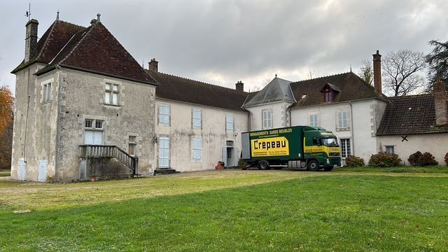 Camion de déménagement blanc circulant dans une rue en ville, avec des bâtiments élevés et le soleil en arrière-plan.