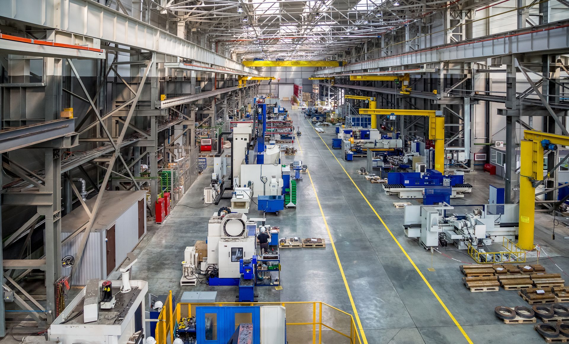Intérieur d’une grande usine industrielle avec des machines, des grues et des postes de travail le long d’une allée centrale.