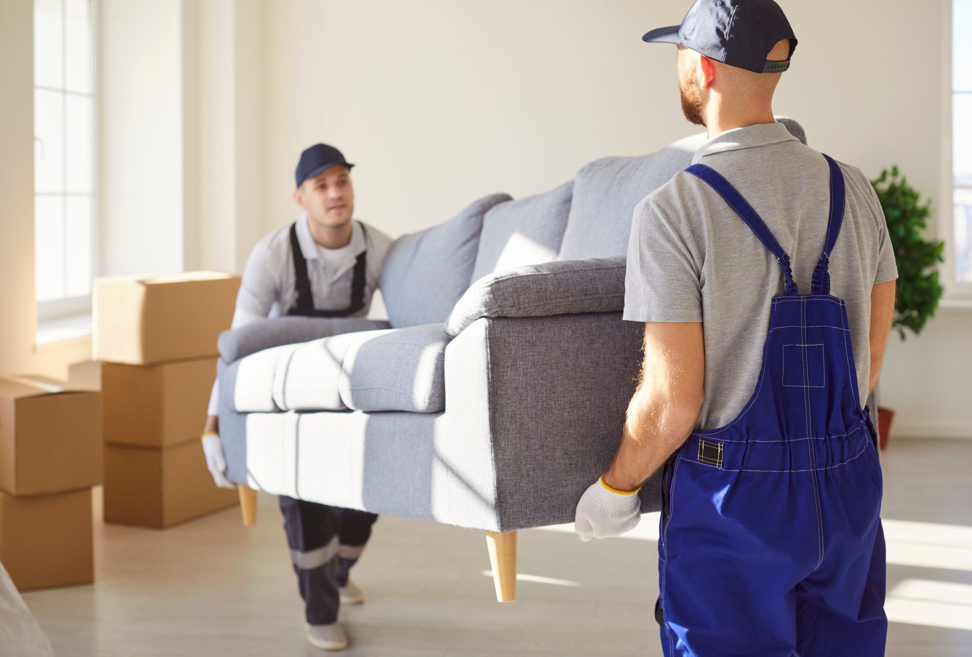 Deux déménageurs portent un canapé gris dans un salon lumineux rempli de cartons de déménagement.