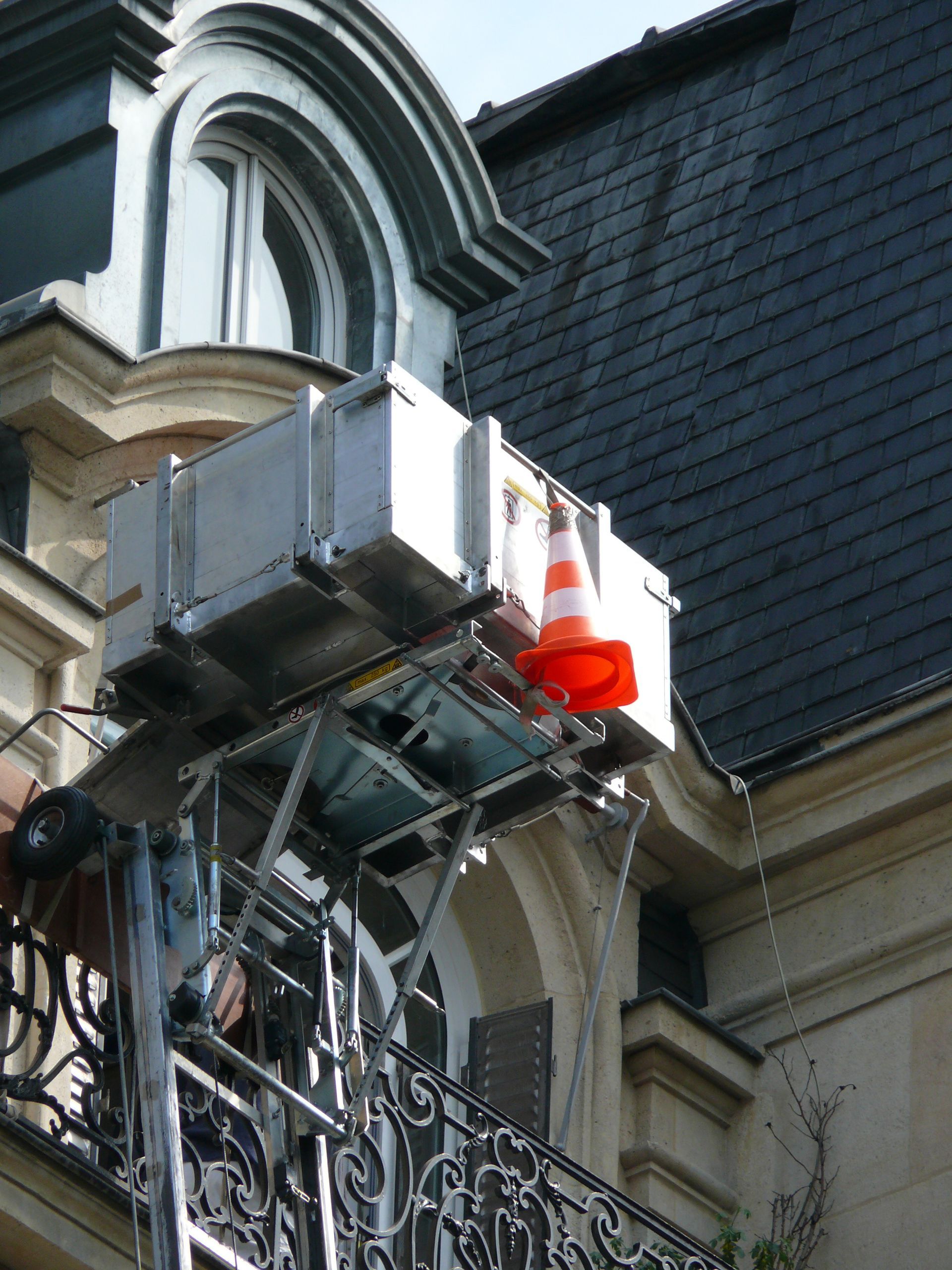 Plateforme utilitaire avec des cônes de signalisation orange fixés à la façade d’un bâtiment, près d’une fenêtre et d’ornements en pierre sculptés.