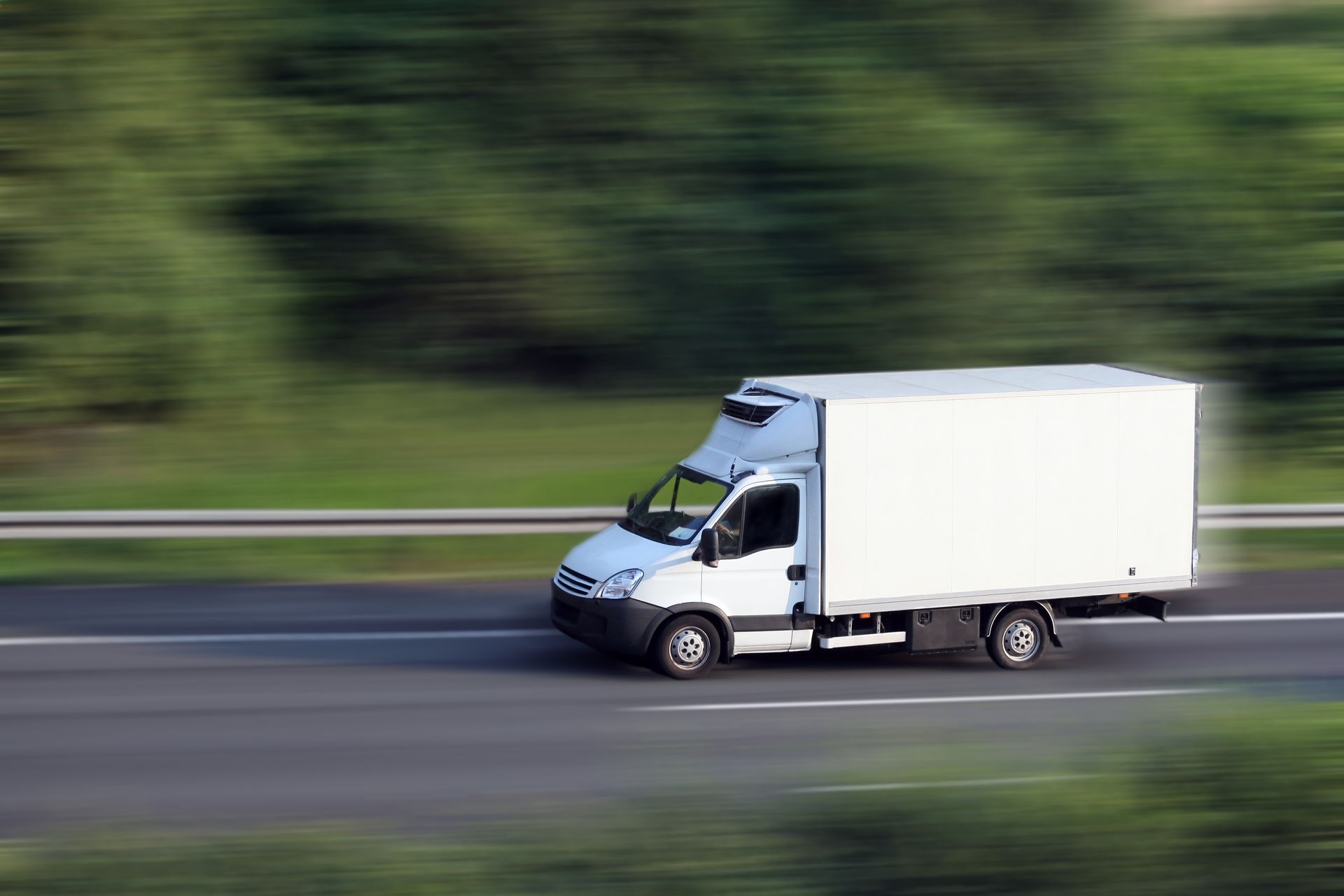 Camion de livraison blanc roulant à vive allure sur une autoroute floue, avec des arbres verts en arrière-plan.