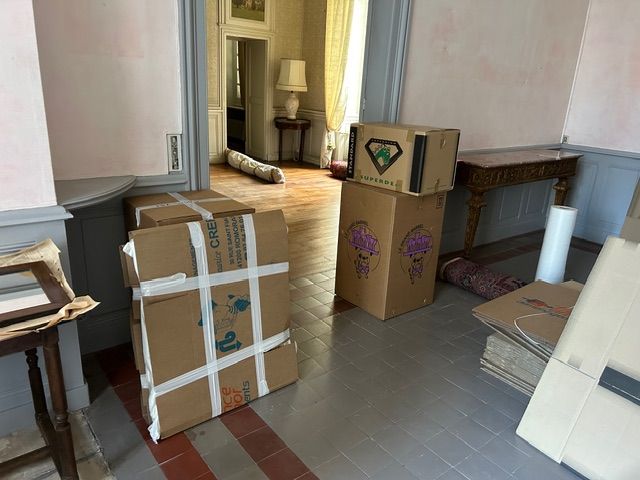 Des cartons de déménagement empilés dans le couloir d’une pièce vide au sol en bois.