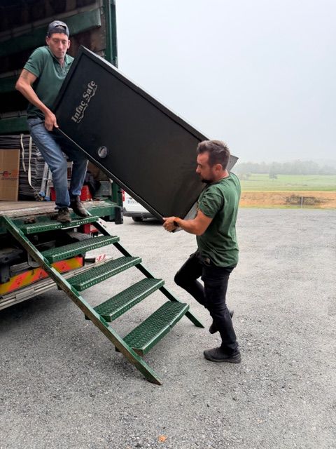 Deux ouvriers chargent une armoire noire dans un camion vert en montant les marches, dans une cour à la campagne