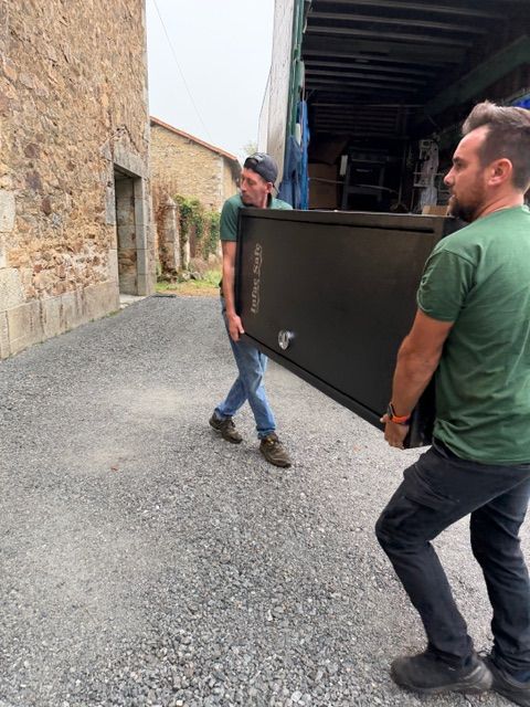 Deux hommes transportent un grand panneau de métal sombre dans une étroite ruelle gravillonnée bordée de bâtiments en pierre.
