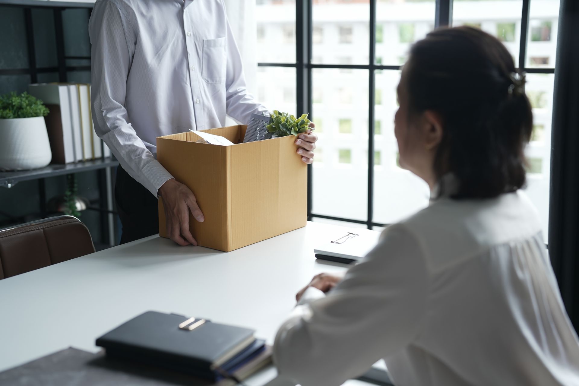 Un homme tient un carton posé sur un bureau, une personne est assise à proximité.