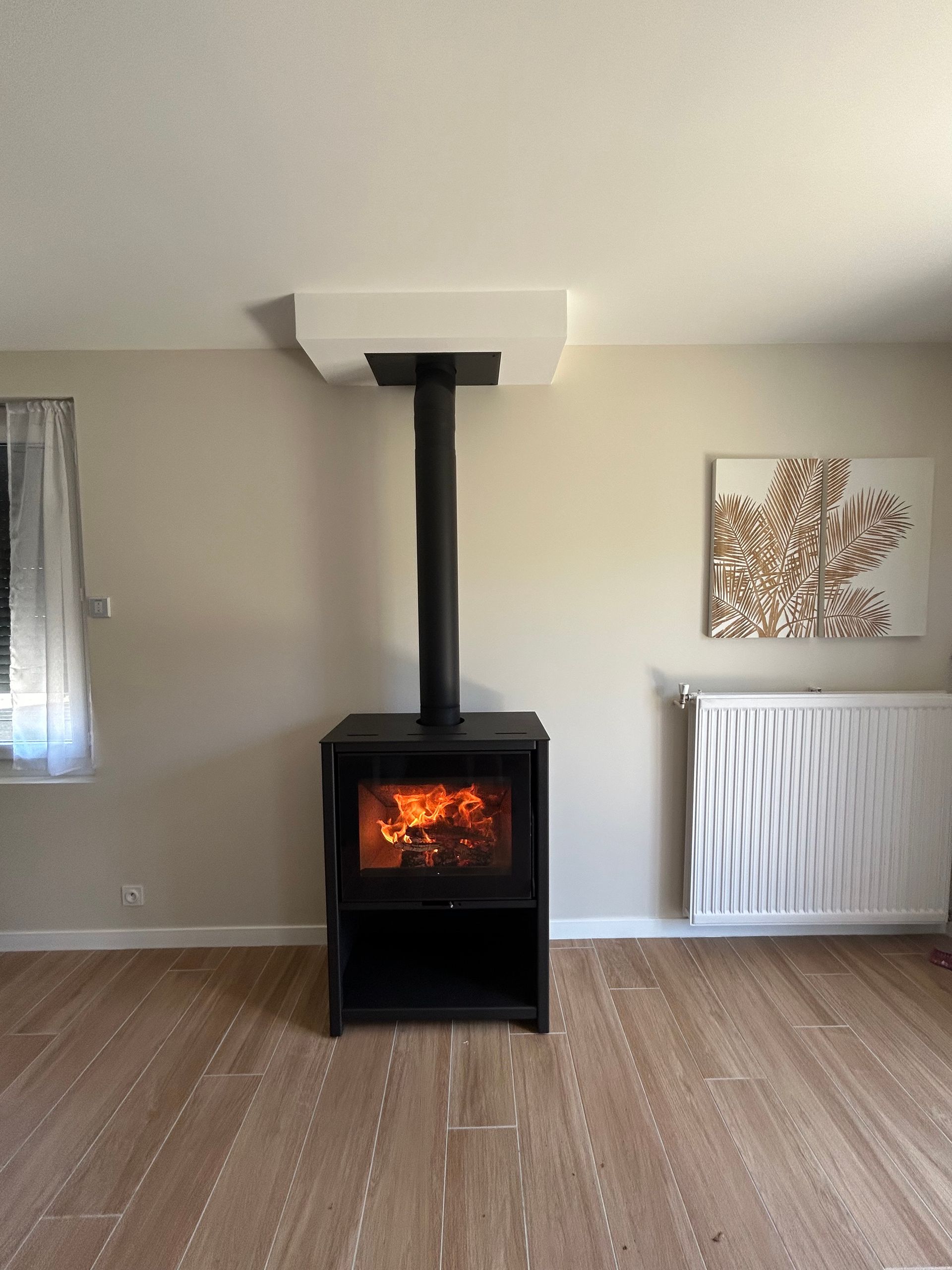 Poêle à bois avec flammes, conduit de cheminée noir, sur plancher de bois, murs beiges, plafond blanc, radiateur blanc et œuvre d'art représentant un arbre