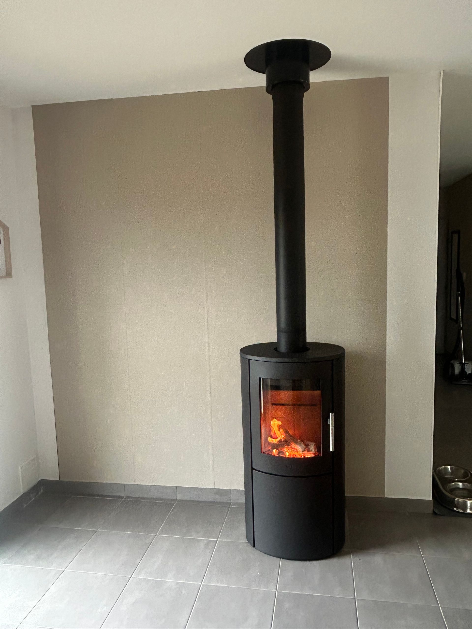 Poêle à bois noir avec feu allumé, cheminée noire, devant un mur marron clair. Sol carrelé gris