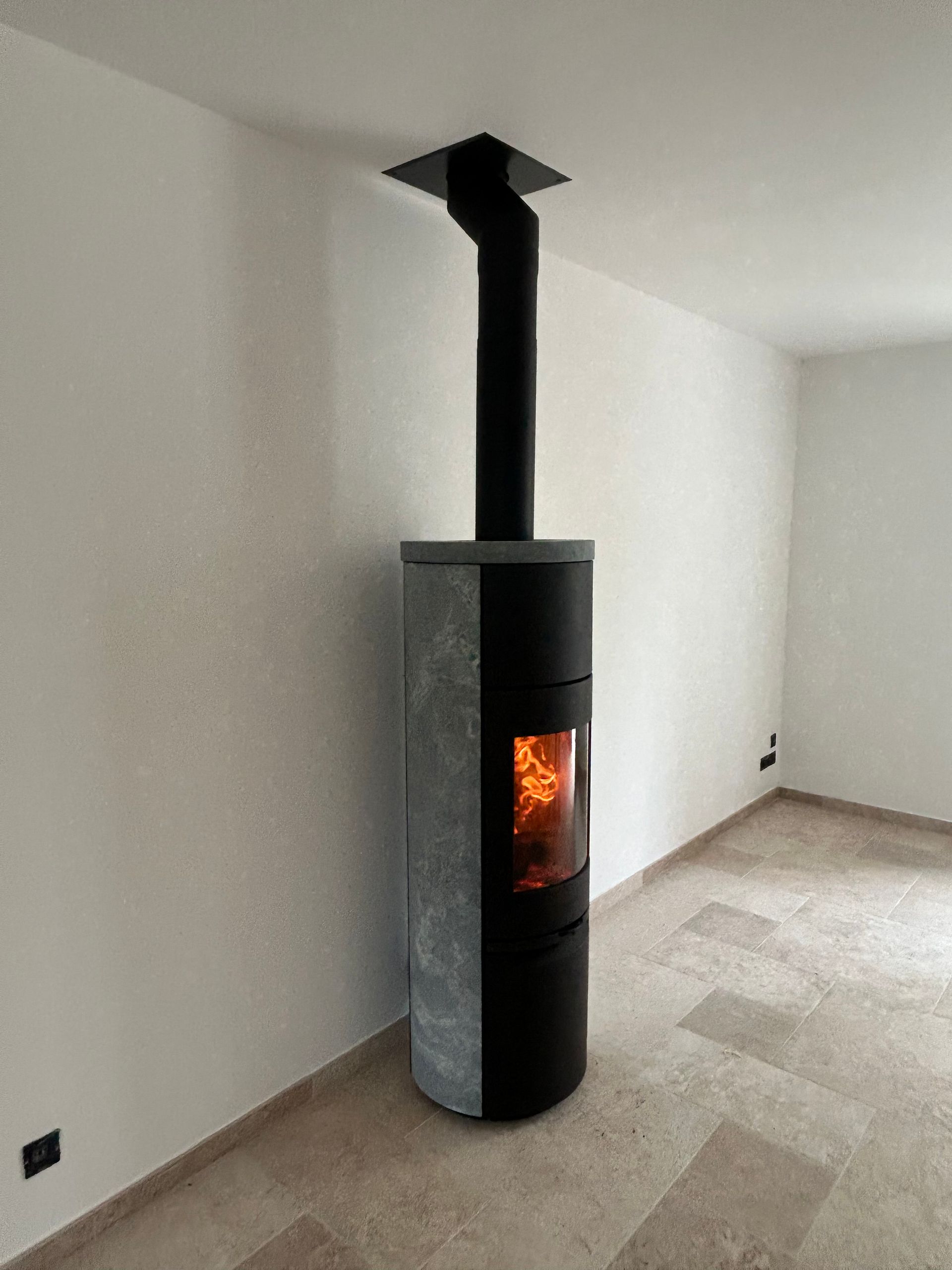 Un poêle à bois moderne avec une cheminée noire montant jusqu'au plafond. Un feu brûle à l'intérieur