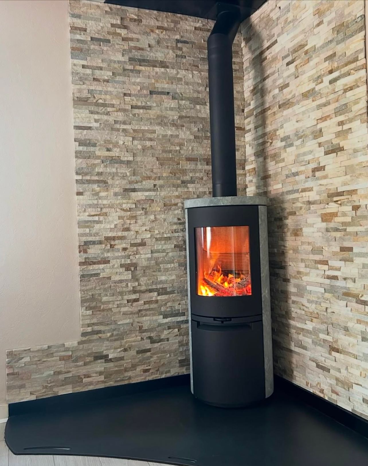 Poêle à bois dans un coin, feu allumé visible. Mur en pierre en arrière-plan