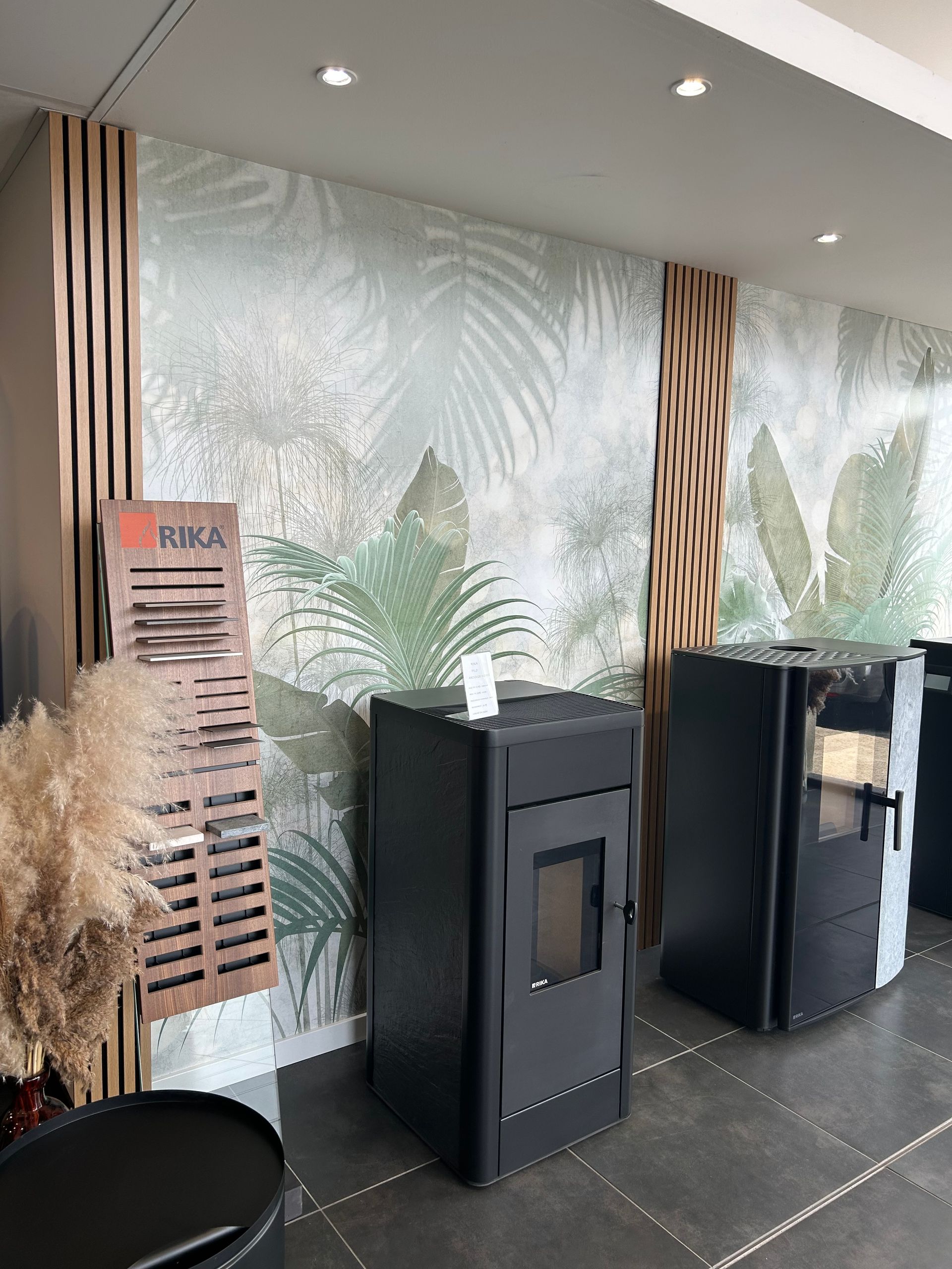 Une salle d'exposition avec des poêles noirs sur fond de fresque tropicale, de boiseries et d'échantillons de produits