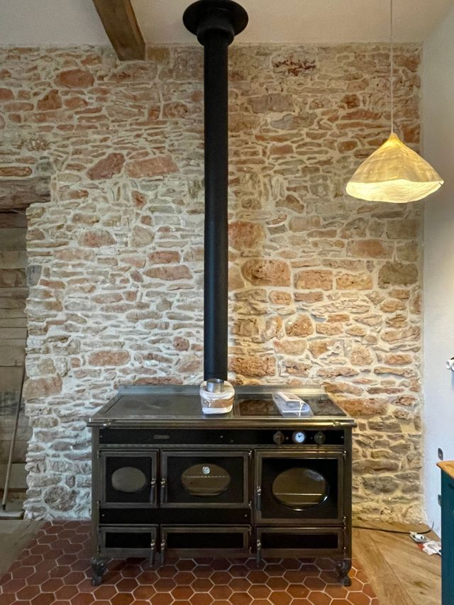 Cuisine aux murs en pierre avec un poêle à bois noir, une cheminée noire et une suspension tissée