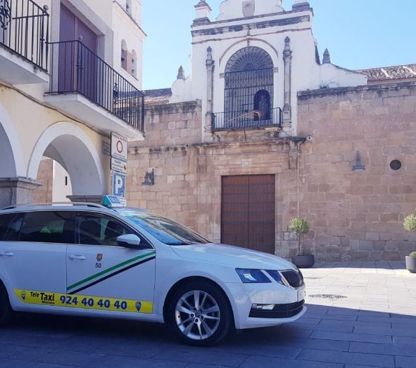 Taxi blanco estacionado frente a una iglesia en una plaza. Letrero de taxi verde y amarillo; edificio de piedra beige.
