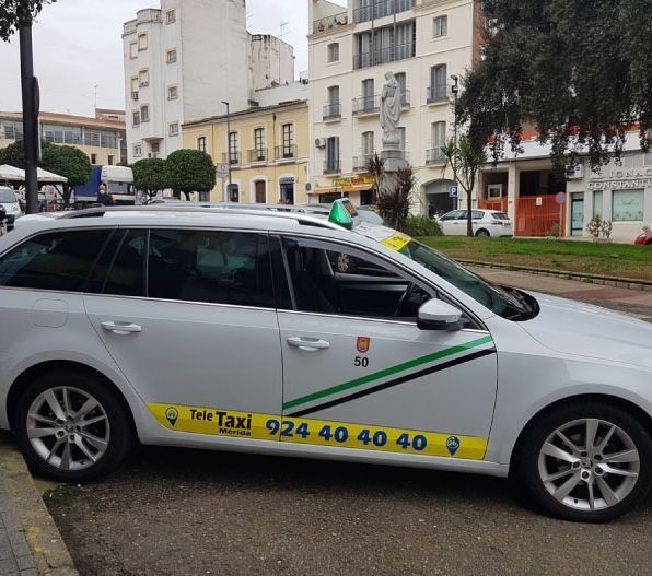 Taxi blanco estacionado al costado de una calle de la ciudad, con una pegatina verde y amarilla y un número de teléfono en el costado.