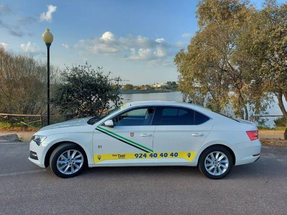 Taxi blanco estacionado en una calle, con marcas verdes y amarillas. Número 924 40 40 40. Lago al fondo.