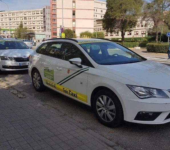 Taxi blanco estacionado en una calle de la ciudad, otros coches y un edificio visibles al fondo.