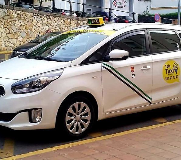 Taxi blanco estacionado con un cartel amarillo en el techo, rayas verdes y negras y número de teléfono en el costado.