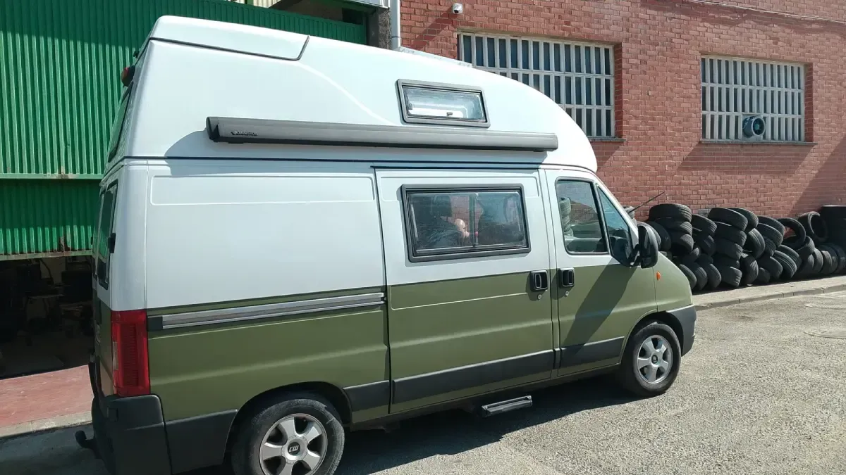 Una autocaravana blanca y verde aparcada junto a un almacén y una pila de neumáticos.
