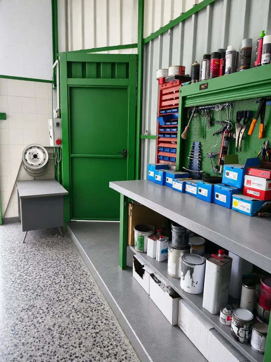 Rincón de taller verde con banco de trabajo, herramientas, estantes y un ventilador de metal junto a la puerta.