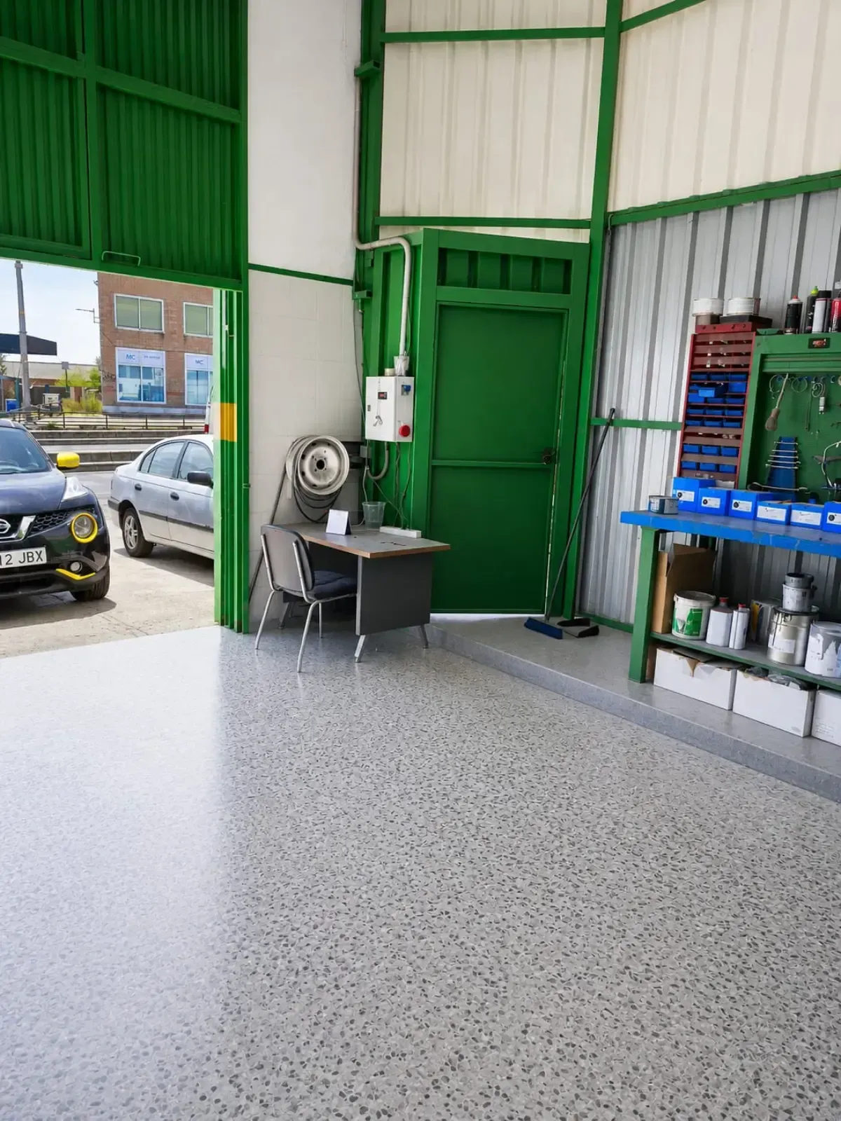 Interior de taller industrial verde con puerta de acceso abierta, banco de trabajo y estanterías metálicas.