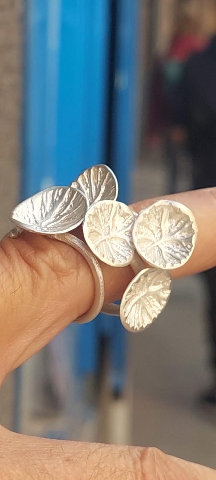 Mano sosteniendo cinco brazaletes plateados con diseño de hojas sobre un fondo azul difuminado.