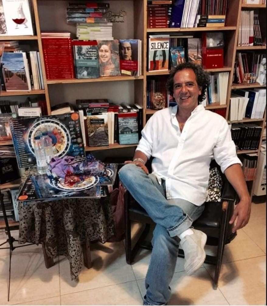 Un hombre sentado en una silla de una librería, sonriendo, con estanterías y coloridas obras de arte detrás de él.