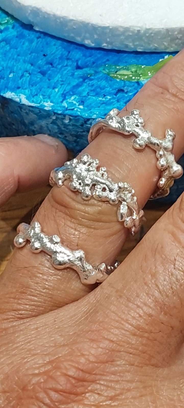 Primer plano de una mano que lleva cuatro anillos de plata con cuentas y piedras transparentes sobre un fondo azul.