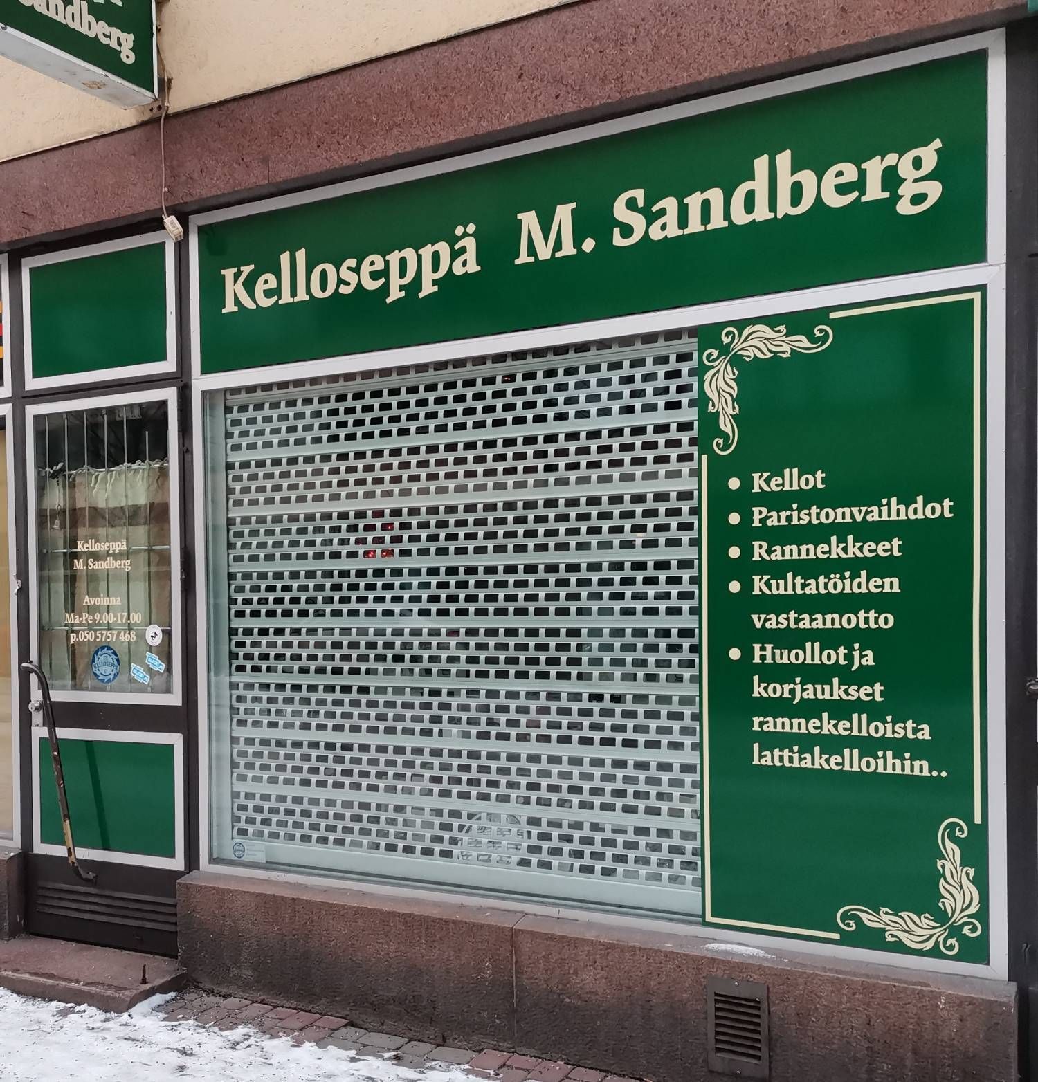Green storefront sign for “Kelloseppä M. Sandberg” with a closed metal shutter and service list.