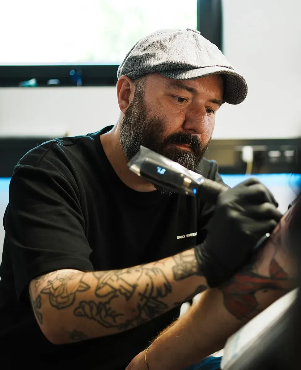 Eine tätowierte Person mit Bart und Kappe, benutzt in einem Studio eine Tätowiermaschine am Arm eines Kunden.