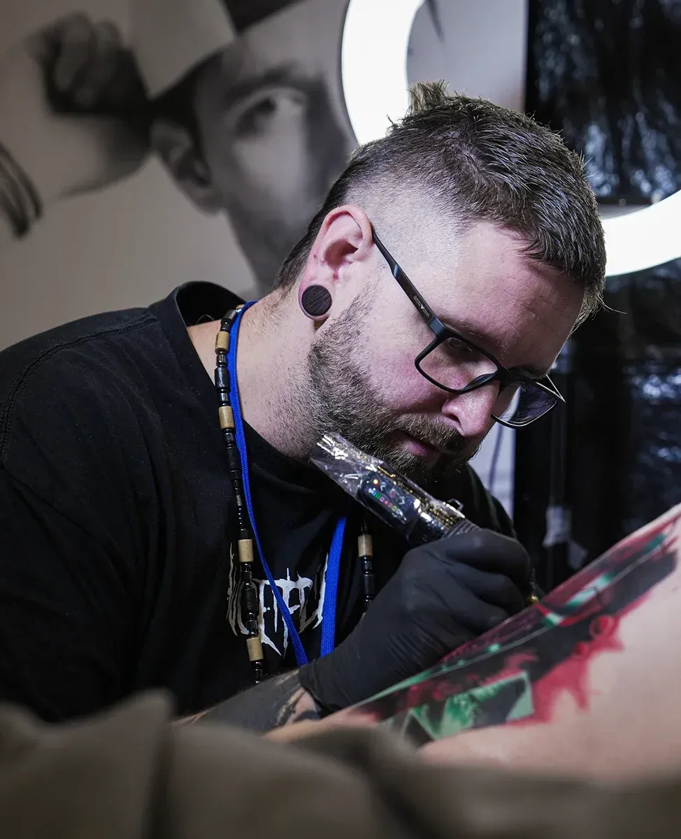 Ein tätowierter Künstler mit Brille und Handschuhen konzentriert sich angestrengt beim Tätowieren der Haut eines Kunden.