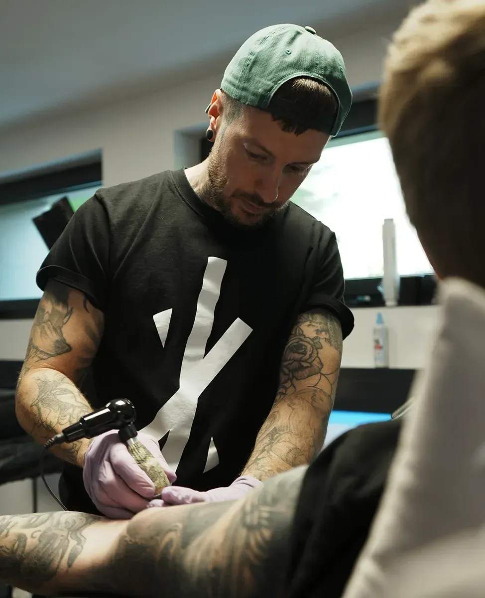 Ein Tätowierer mit grüner Kappe und schwarzem Hemd konzentriert sich angestrengt beim Tätowieren des Arms eines Kunden.