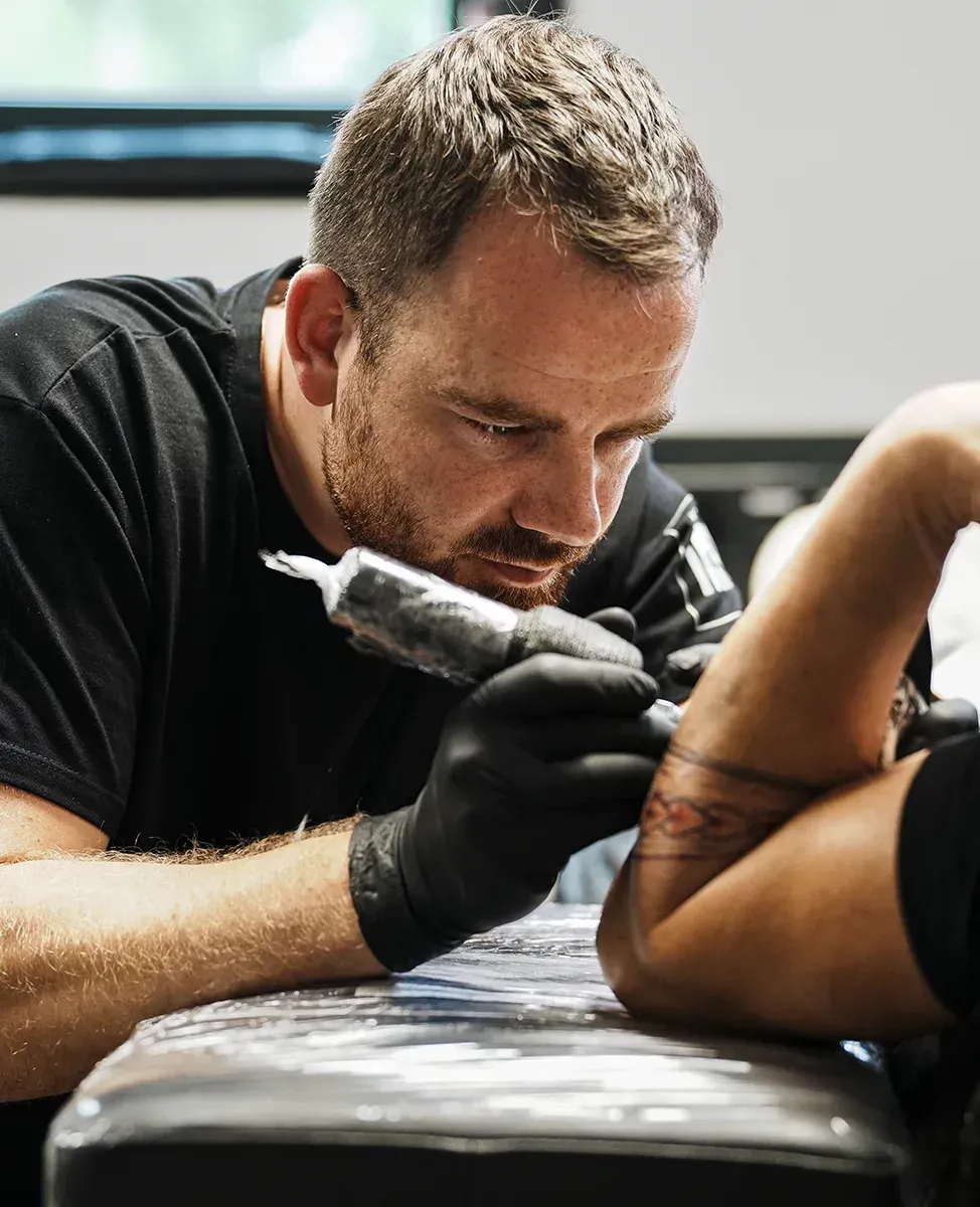 Ein Tätowierer in schwarzem T-Shirt und Handschuhen trägt sorgfältig Tinte auf den Arm eines Kunden auf.