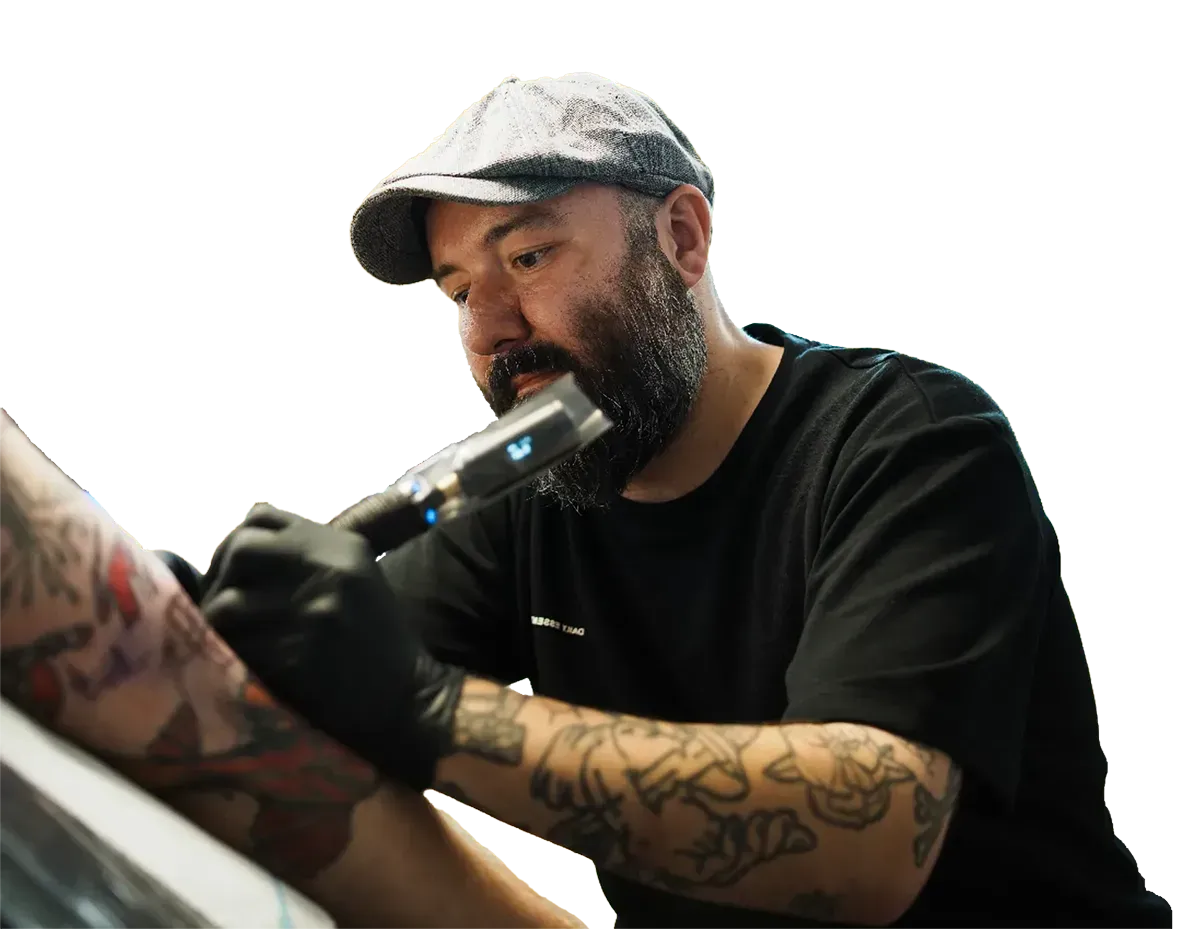 Eine bärtige Person mit Schiebermütze und schwarzen Handschuhen benutzt eine Tätowiermaschine am Arm eines Kunden.