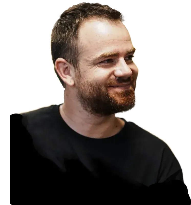 Eine Person mit kurzen, dunklen Haaren und Bart, die ein schwarzes Hemd trägt und mit einem sanften Lächeln zur Seite blickt.