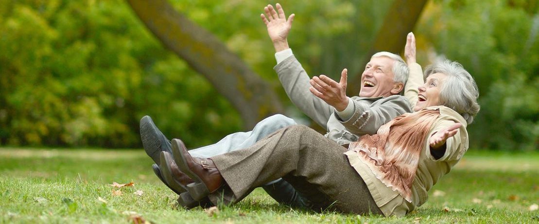 Una pareja de ancianos riendo y levantando los brazos mientras están acostados en un parque con césped.