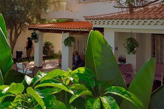 El follaje verde enmarca un edificio blanco con techo de tejas rojas; la gente se sienta y se para en el porche.