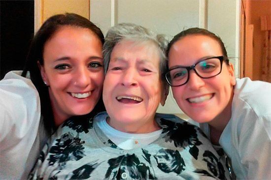 Mujer con dos mujeres más jóvenes, todas sonriendo. Interior, colores claros.
