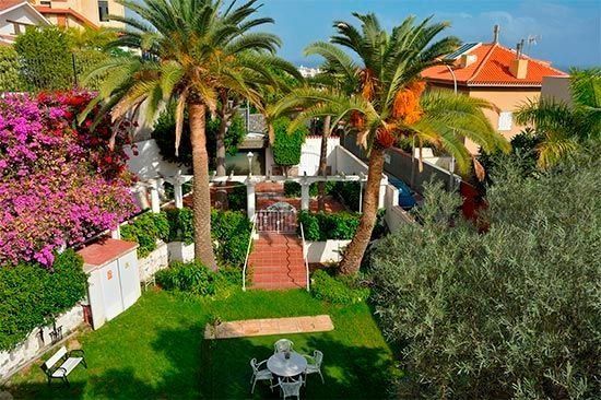 Exuberante jardín verde con palmeras, camino de ladrillos rojos, cenador blanco y una mesa y sillas.