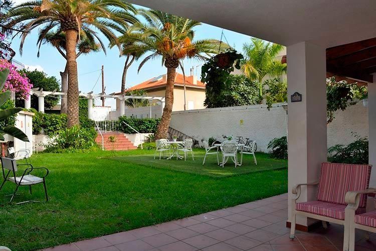 Césped con palmeras, mesas y sillas blancas y un patio con sillas bajo un alero.