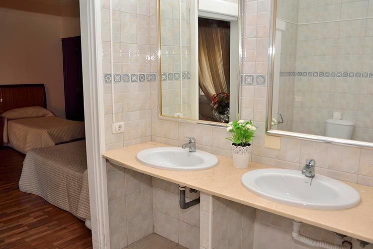 Baño con dos lavabos, espejo y una vista de dos camas al fondo.