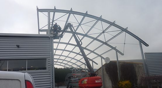 Des ouvriers du bâtiment, sur une nacelle élévatrice, assemblent une charpente métallique incurvée à côté d'un bâtiment.