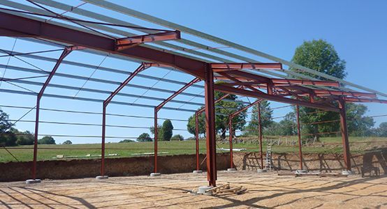 La structure métallique d'un bâtiment en construction, les poutres rouges, le ciel bleu et un champ herbeux.